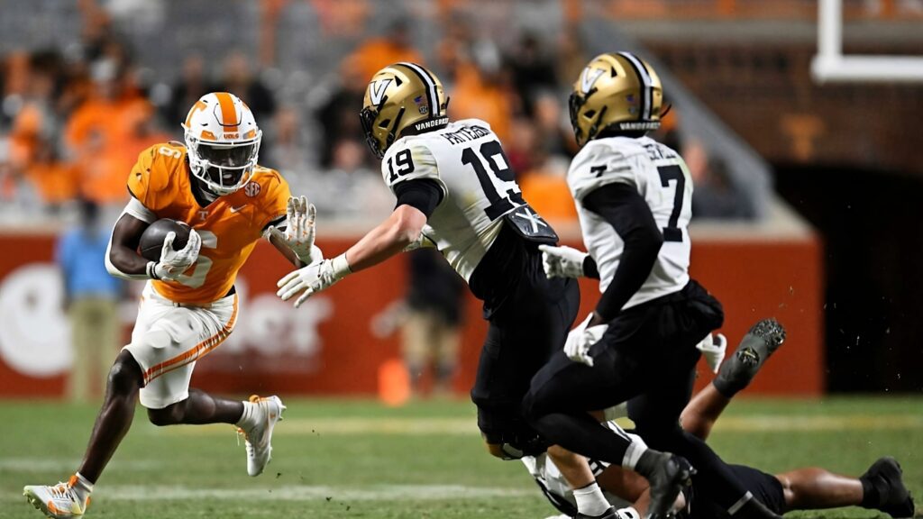 A Tennessee Volunteers football player in an orange jersey runs with the ball, pursued by two Vanderbilt Commodores players in white and black jerseys during a game