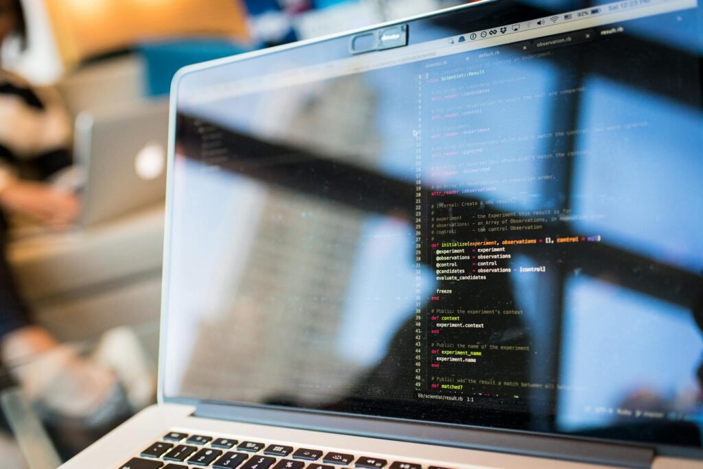 A close-up of a laptop screen displaying code with a blurred background of a person using another laptop